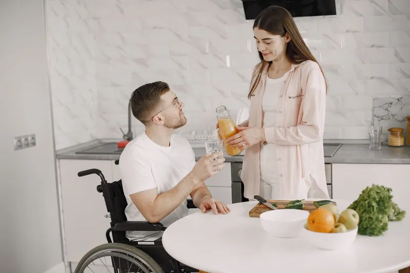 A young man who uses a wheelchair smiles as a woman pours him a glass of juice in a bright home kitchen.