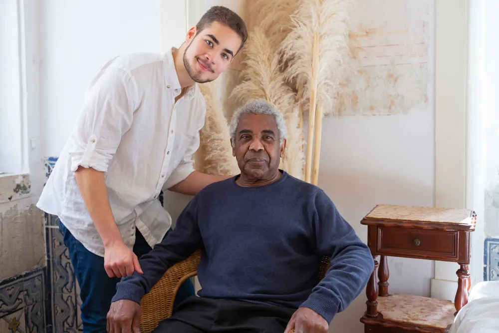 A young man in a white shirt smiles as he stands beside an older man seated in a wicker chair in a sunny living room.